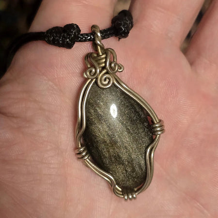 Ciondolo artigianale in ossidiana dorata con montatura in filo metallico argentato tenuto in mano.